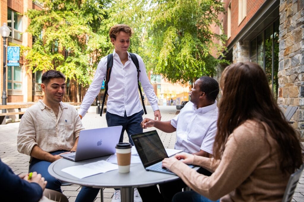 Three students study together outside and another stops to talk to them.